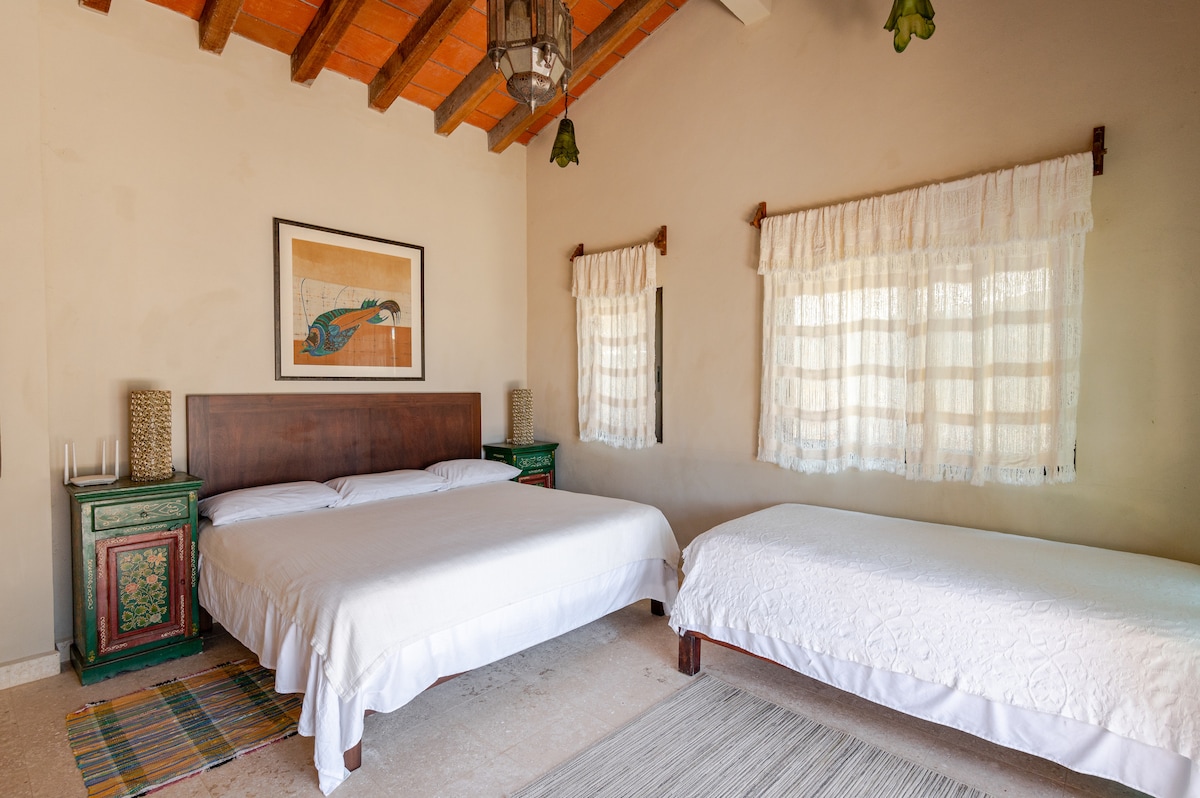 A spacious sleeping area is furnished with a comfortable queen size bed and a single bed. Natural light filters through the window treatments, highlighting the antique nightstands and vibrant wall art. The neutral tones create a calming environment.