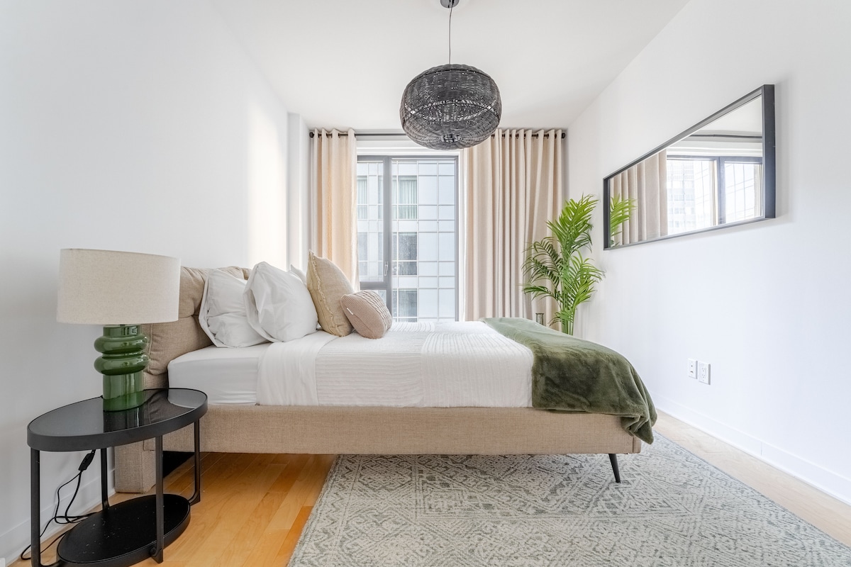 A well-appointed bedroom features a spacious bed adorned with neutral-toned pillows and a soft green throw. Natural light enters through a large window with sheer curtains. A decorative mirror enhances the space, while a small, modern lamp and a potted plant add touches of interest.