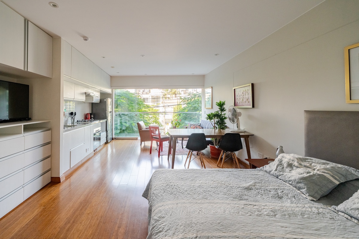 A spacious loft features a comfortable sleeping area with a soft gray bedspread. The modern furnishings include a dining table surrounded by chairs and a kitchen area equipped with cabinetry and appliances. Large glass doors open to a balcony overlooking lush greenery.