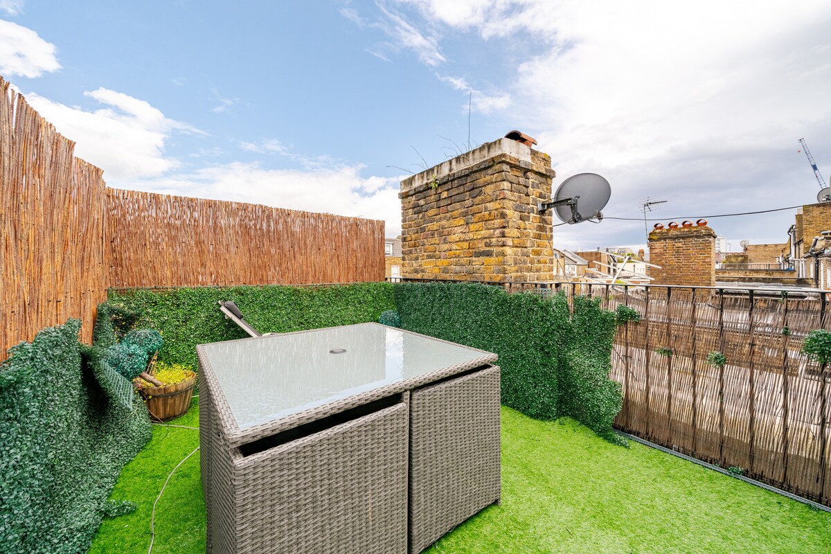 A private outdoor terrace features a glass-top table surrounded by cushioned seating. Lush green landscaping and a natural grass surface create a serene atmosphere. The view includes a skyline framed by a brick chimney and nearby rooftops, enhancing the space's tranquility.