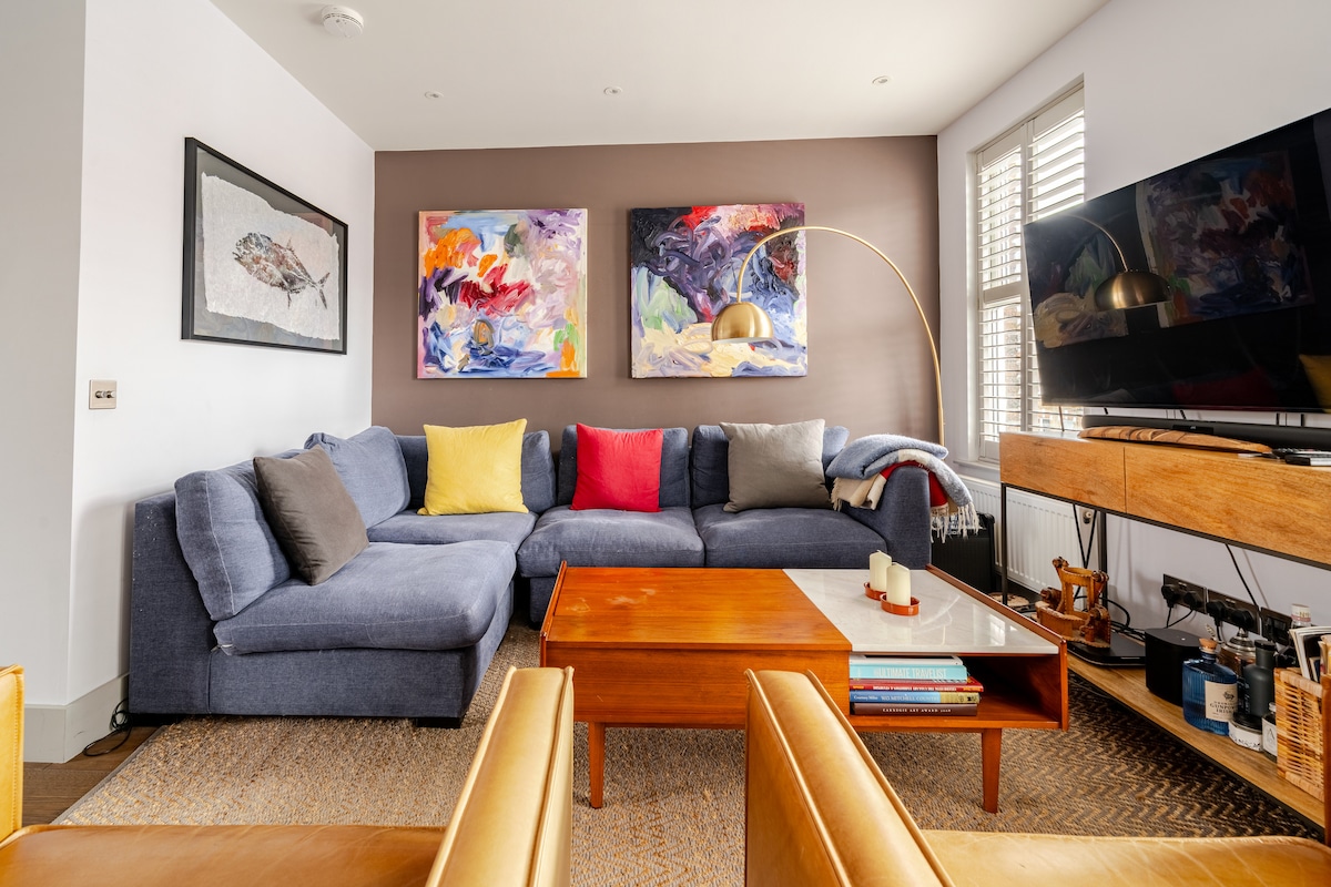 A spacious living area features a large sectional sofa adorned with colorful cushions, complemented by two vibrant abstract paintings on the wall. A wooden coffee table sits at the center, surrounded by sleek armchairs, and a modern television is positioned on a wooden console.