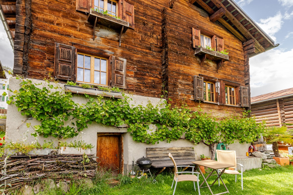 The exterior of a charming wooden house is shown, featuring traditional shutters and climbing greenery. A small table and chairs are set on the grassy area, with a barbecue nearby. The structure reflects rustic alpine architecture, complemented by a serene, natural landscape.