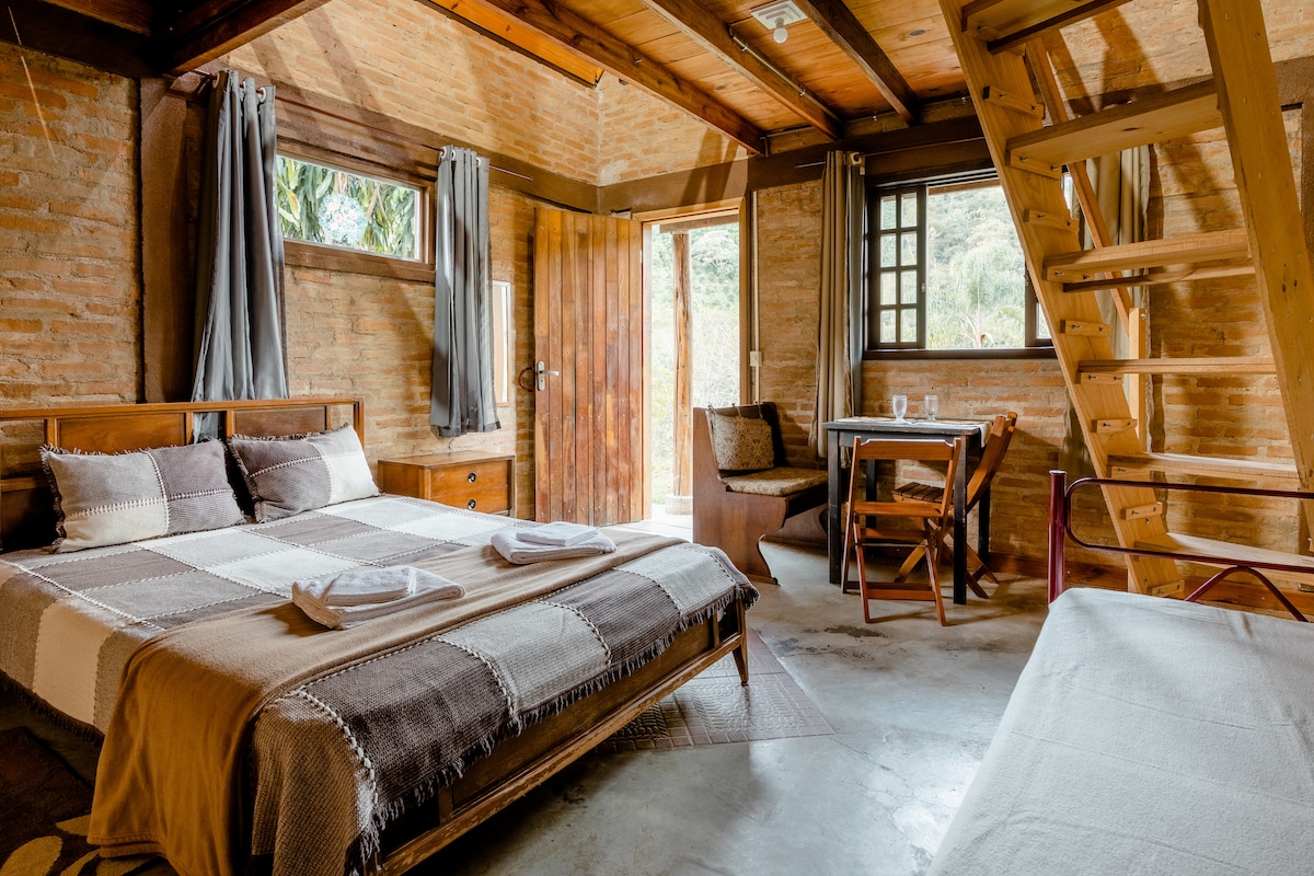 A rustic interior features a spacious bed adorned with patterned bedding. A wooden table and chairs are positioned nearby, allowing for dining. Natural light enters through the window, while a wooden staircase leads to a loft area.