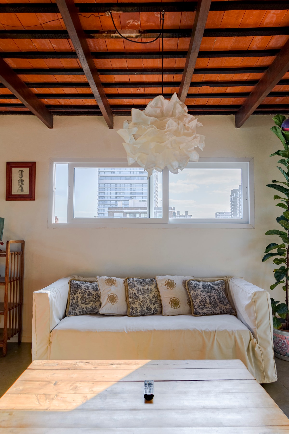 A cozy living space features a white couch adorned with decorative pillows. A wooden coffee table occupies the center, complemented by a remote control. Natural light enters through large windows framed by tall green plants, enhancing the airy ambiance of the room.