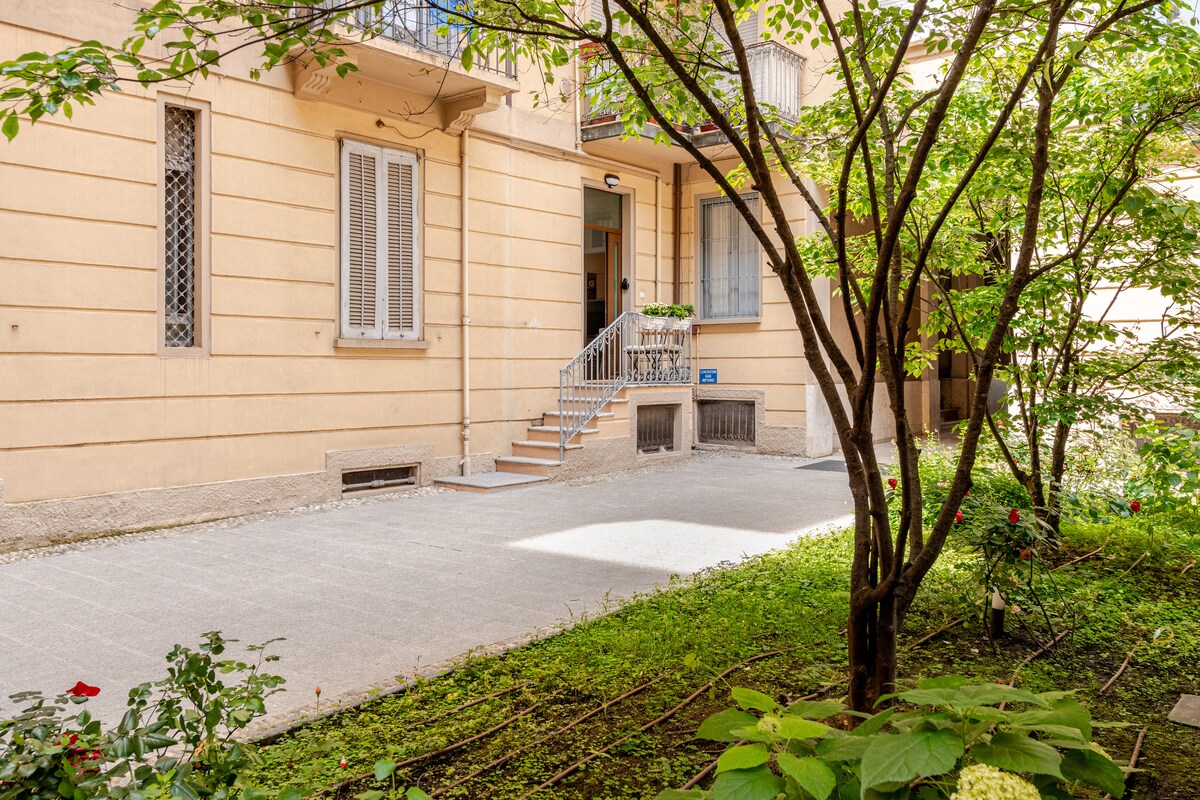 A serene courtyard is framed by lush greenery and well-maintained plants. A set of stairs leads to a welcoming entrance, complemented by soft natural light filtering through the trees. The courtyard's paved area provides ample space for relaxation.
