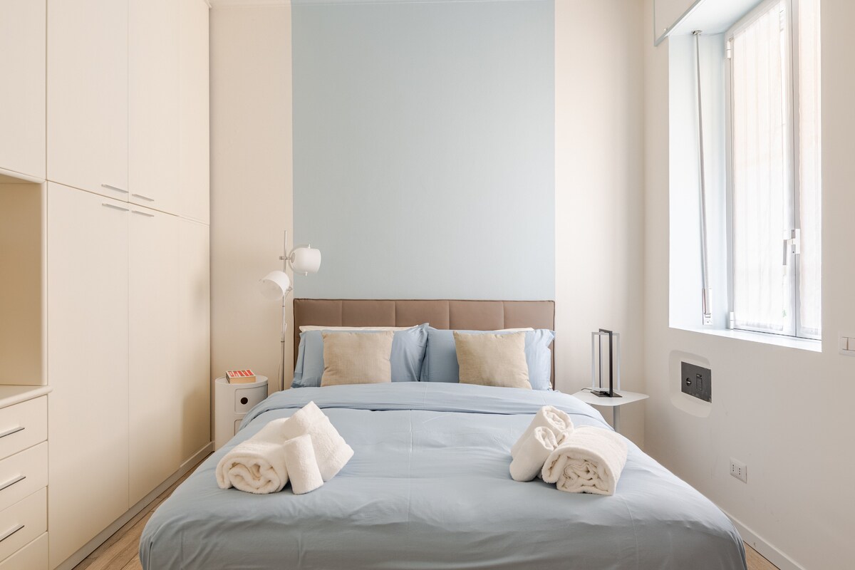 A well-appointed bedroom is highlighted by a spacious bed with a light blue comforter. Two neatly folded towels are displayed at the foot of the bed. A bedside table complements the decor, with soft natural light streaming through a window adorned with light curtains.