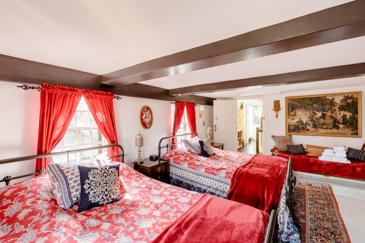 A spacious guest room features two full-size beds adorned with vibrant red and blue bedding. A day bed is positioned against the wall, complemented by an artwork depicting a historical scene. Large windows, draped with bright red curtains, allow natural light to illuminate the room.