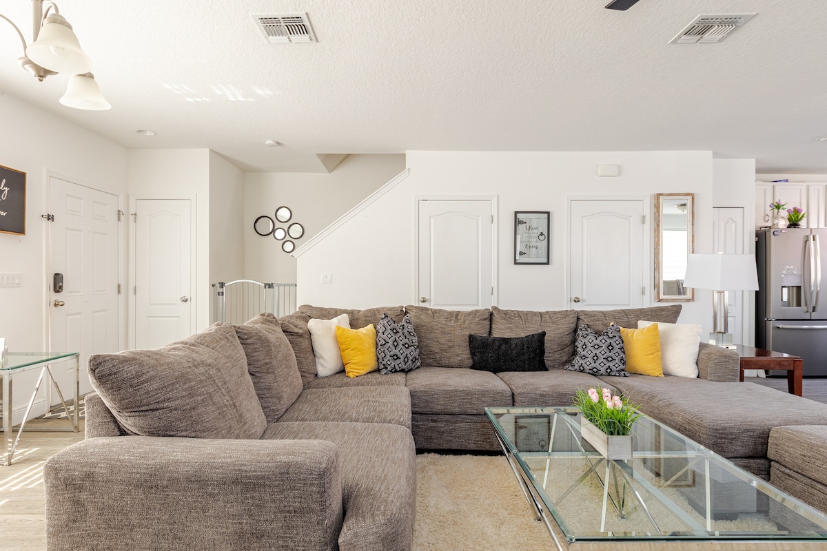 A spacious living area is showcased with a large sectional sofa adorned with decorative cushions. A coffee table is positioned in the center, while soft natural light filters through the window. The room features light-colored walls and a staircase is visible in the background.