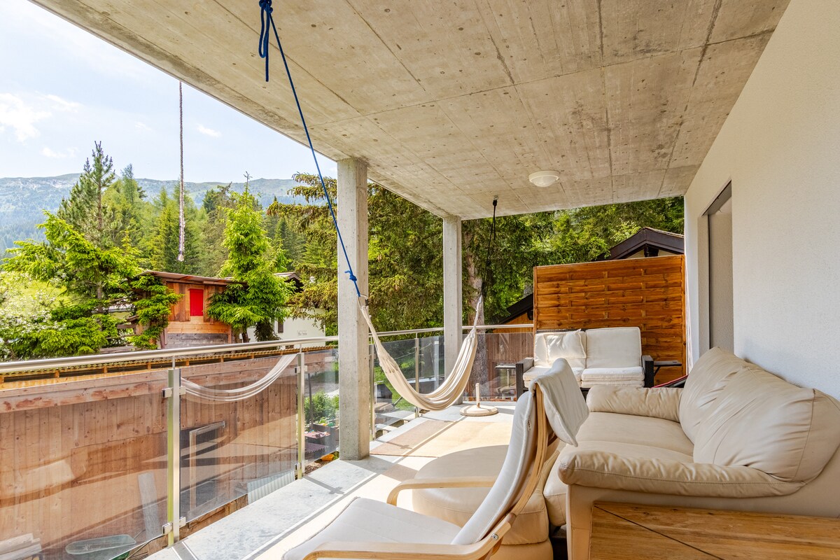 A spacious balcony is furnished with a comfortable sofa and a hammock, providing a relaxed outdoor space. The surrounding greenery and distant mountains create a serene backdrop, with partial views of nearby buildings enhancing the natural setting.