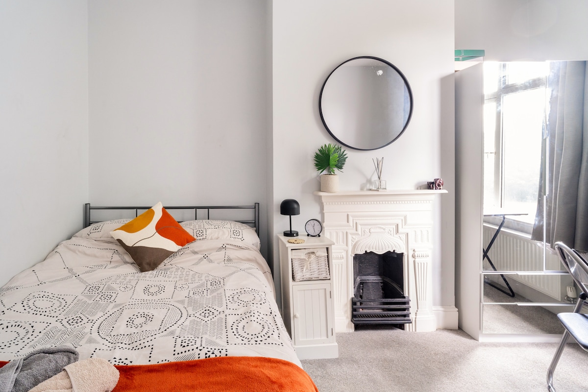 A spacious private room is presented with a double bed featuring a patterned blanket and bright decorative pillows. A round mirror is mounted above a decorative fireplace, while a small desk and chair are placed near a window, providing natural light.