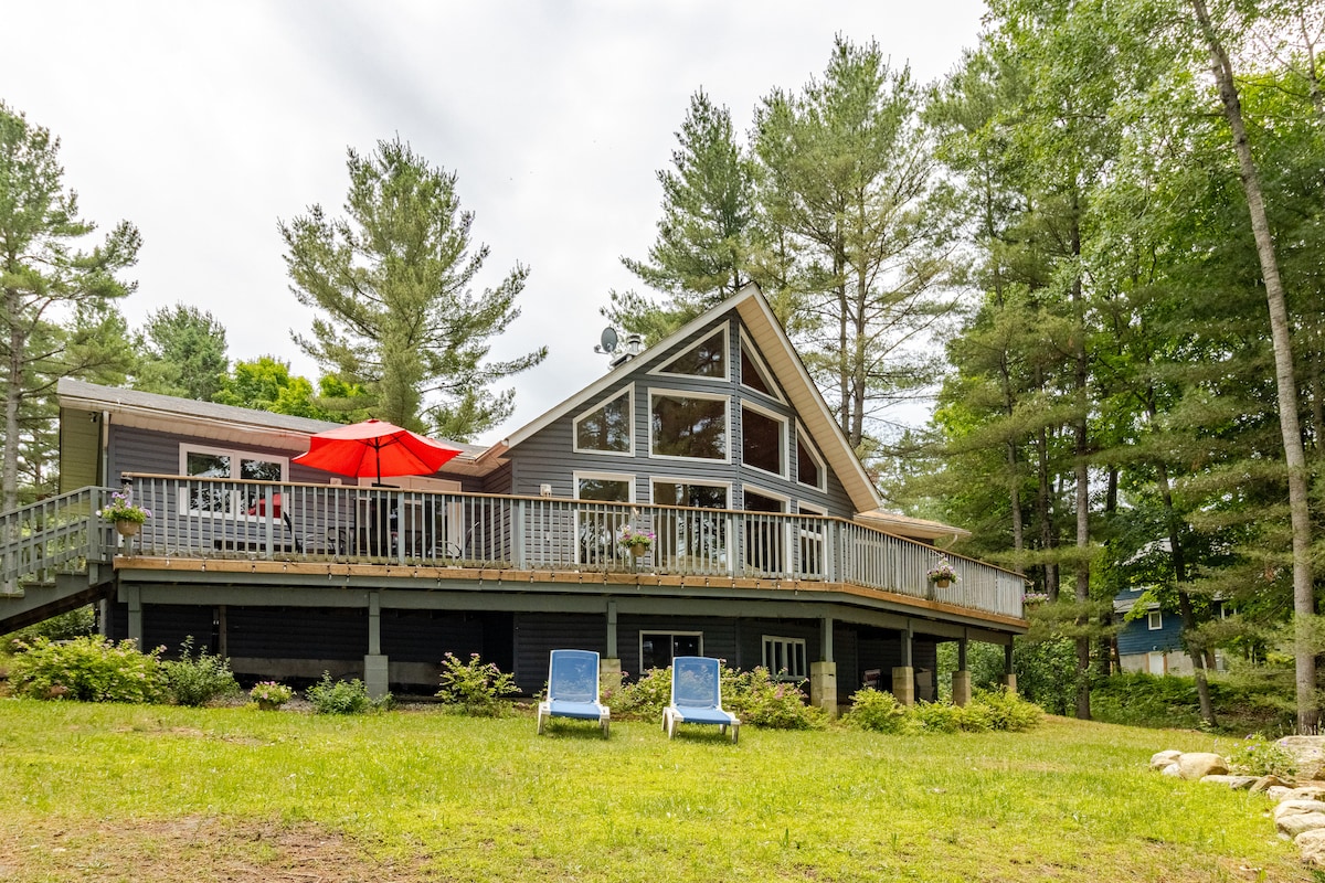 The cottage exterior is shown with a sloped roof and large windows, offering abundant natural light. A spacious deck surrounds the cottage, featuring a bright red umbrella over outdoor seating. Two lounge chairs are positioned on the lawn, with greenery and tall trees in the background.