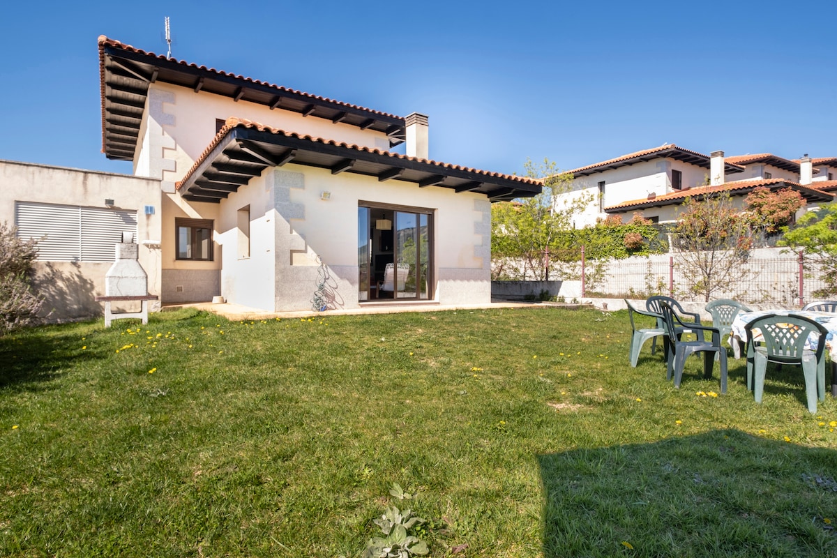 A chalet is presented with a well-maintained garden area surrounded by green grass. Outdoor seating is arranged with several chairs, while the exterior features a mix of light-colored walls and a tiled roof. Large windows offer views of the garden and natural light into the home.