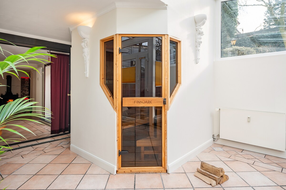 A wooden door with glass panels leads into a sauna, framed by a hexagonal structure. The entrance features a simple logo and is surrounded by light-colored walls and tiled flooring. Potted greenery adds a touch of nature to the welcoming space.