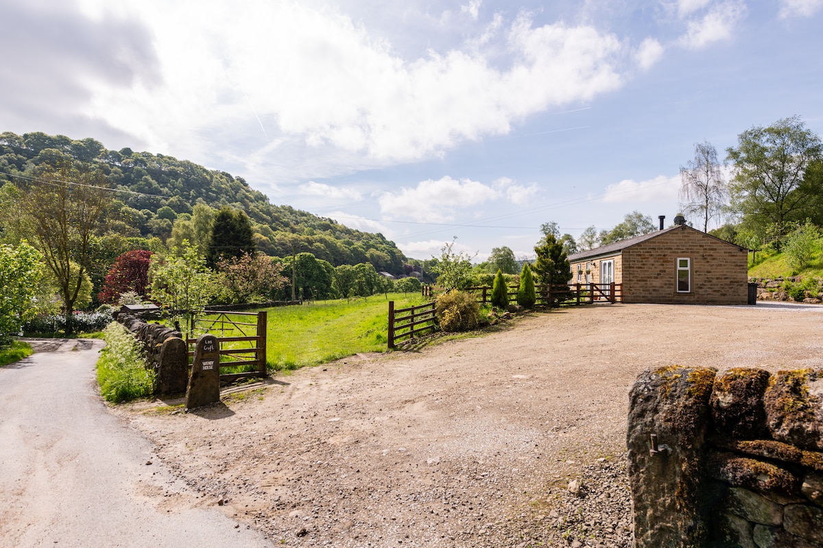The exterior of a semi-detached bungalow is shown, positioned amidst a rural landscape. A gravel parking area is visible, with green fields and trees in the background. The entrance features a wooden gate, leading to the inviting surroundings of the Lee Wood Estate.