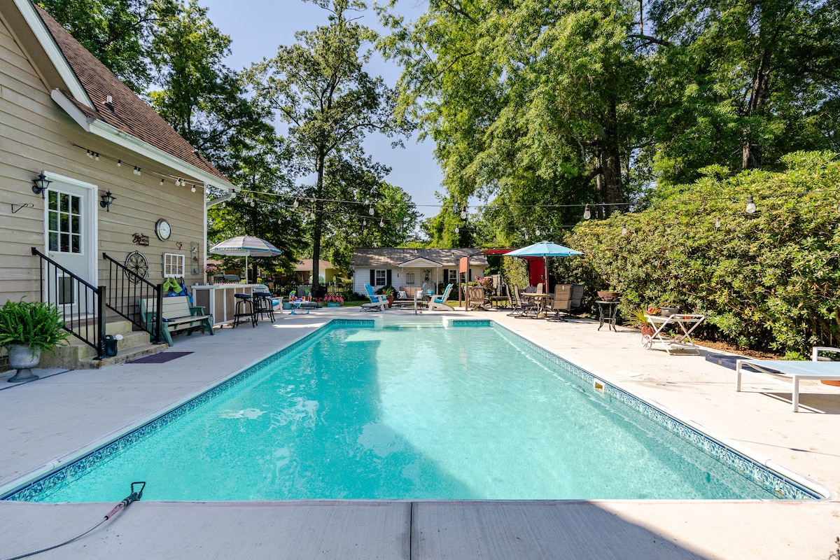A spacious swimming pool is surrounded by a sunlit deck, featuring loungers and shaded seating areas. A combination of vibrant umbrellas offers cool shaded spots. Lush greenery borders the space, creating a tranquil outdoor environment. The inviting blue water of the pool reflects the clear sky.