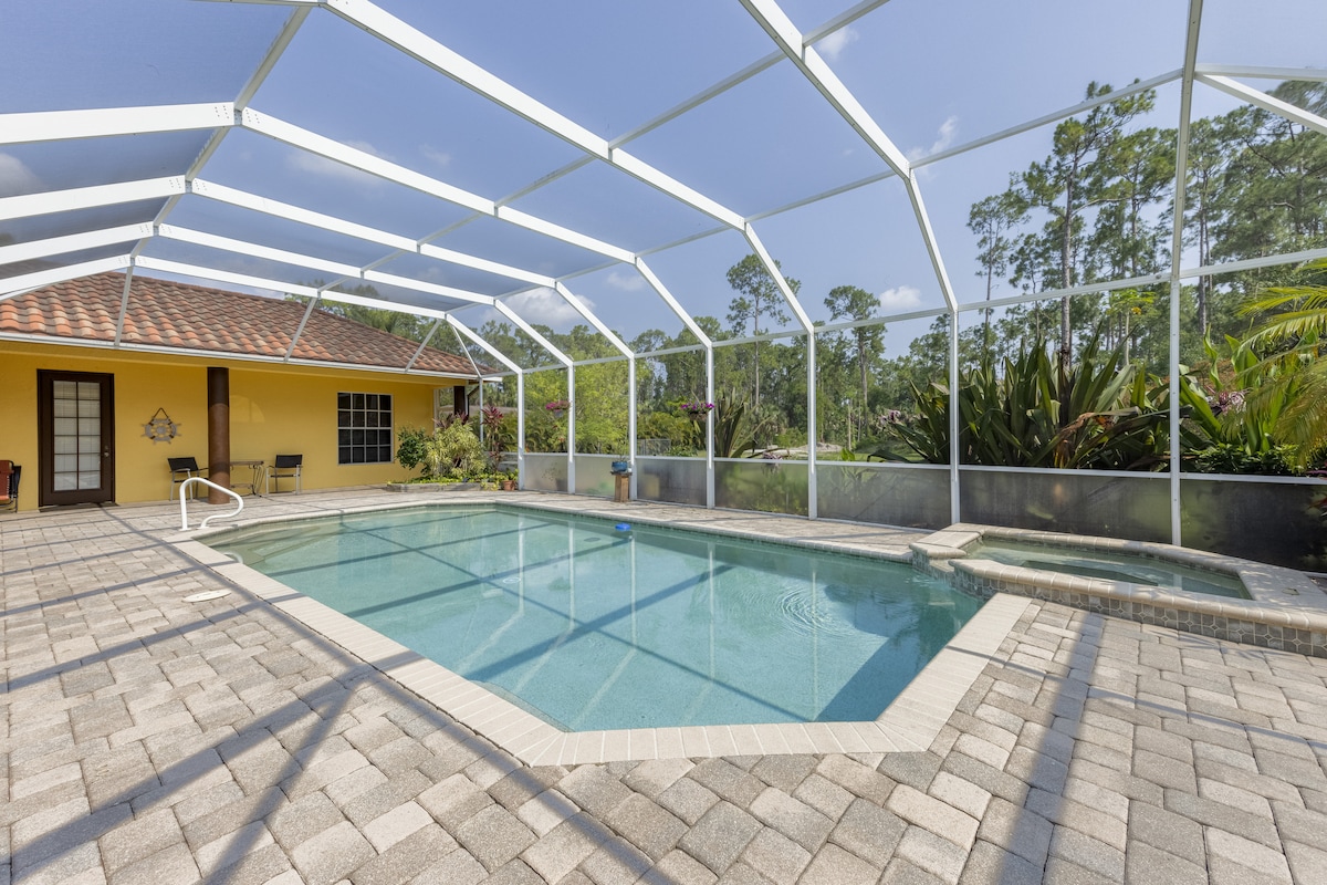 A large, screened lanai encases a bright pool area, featuring a turquoise pool with a surrounding paved deck. Windows offer views of lush greenery and flowering plants, while a doorway leads to an inviting indoor space in the background.