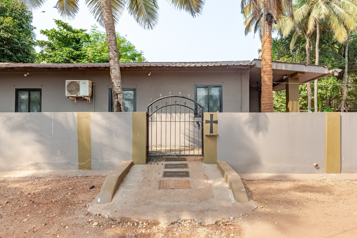 The entrance to the villa is framed by a sturdy gate enclosed by a low wall. Lush palm trees create a serene atmosphere. A simple pathway leads to the door, allowing easy access to the modern structure beyond.
