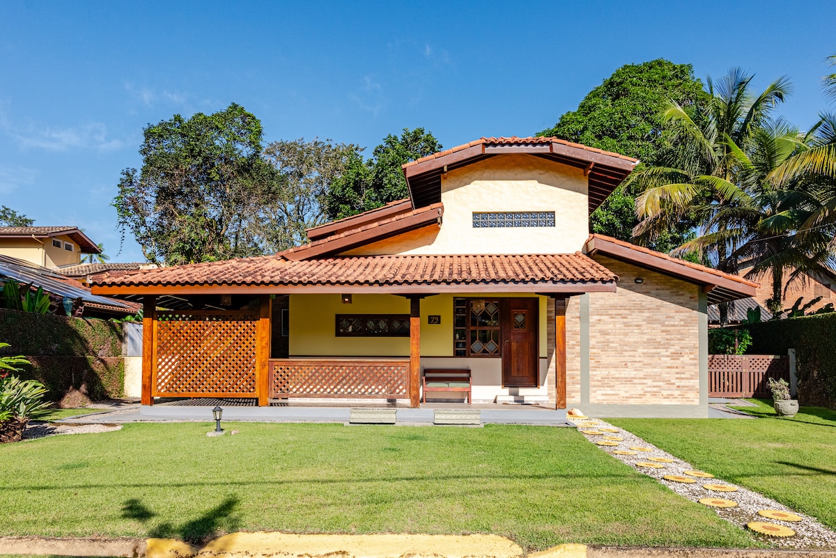The exterior of the house showcases a traditional design, featuring a sloped roof and wooden accents. A well-maintained garden with green grass surrounds the entrance, leading to a covered patio area. Palm trees and foliage create a lush backdrop, emphasizing a tranquil setting.