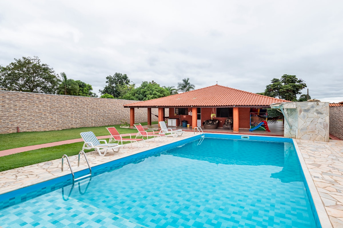 An expansive outdoor area features a large swimming pool surrounded by lounge chairs. A sunlit, roofed space is visible in the background, offering seating and a view of the lush greenery. Well-maintained stone paving enhances the pleasant atmosphere.