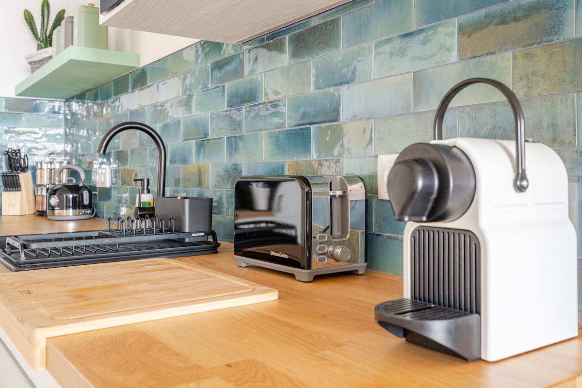 A well-equipped kitchen countertop is displayed, featuring a coffee machine, a toaster, and a black dish rack. The backsplash consists of blue-green tiles, complementing the wooden surface. Kitchen utensils and a cutting board are neatly arranged, providing a functional space for meal preparation.