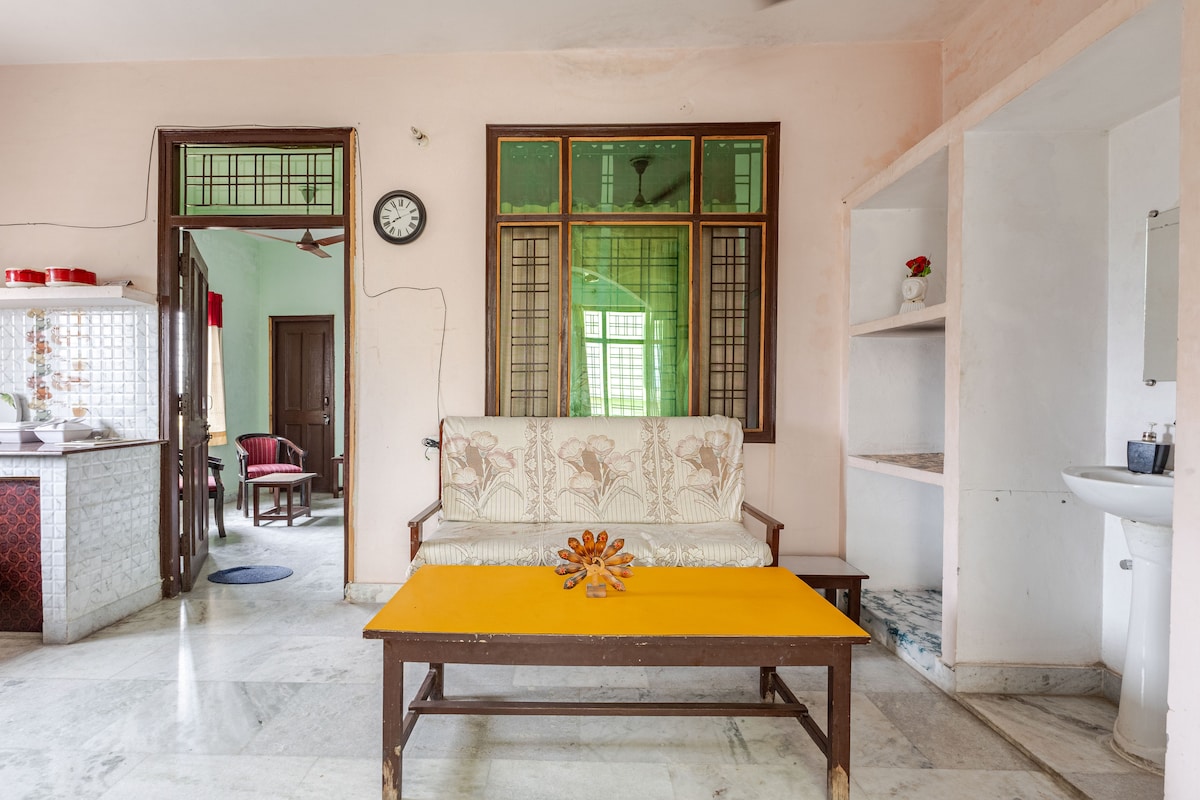 A common lounge area features a light-colored sofa with decorative floral patterns, accompanied by a bright yellow coffee table. Windows allow natural light to fill the room, illuminated against soft pastel walls. A clock and wall-mounted shelf add functional details to the space.