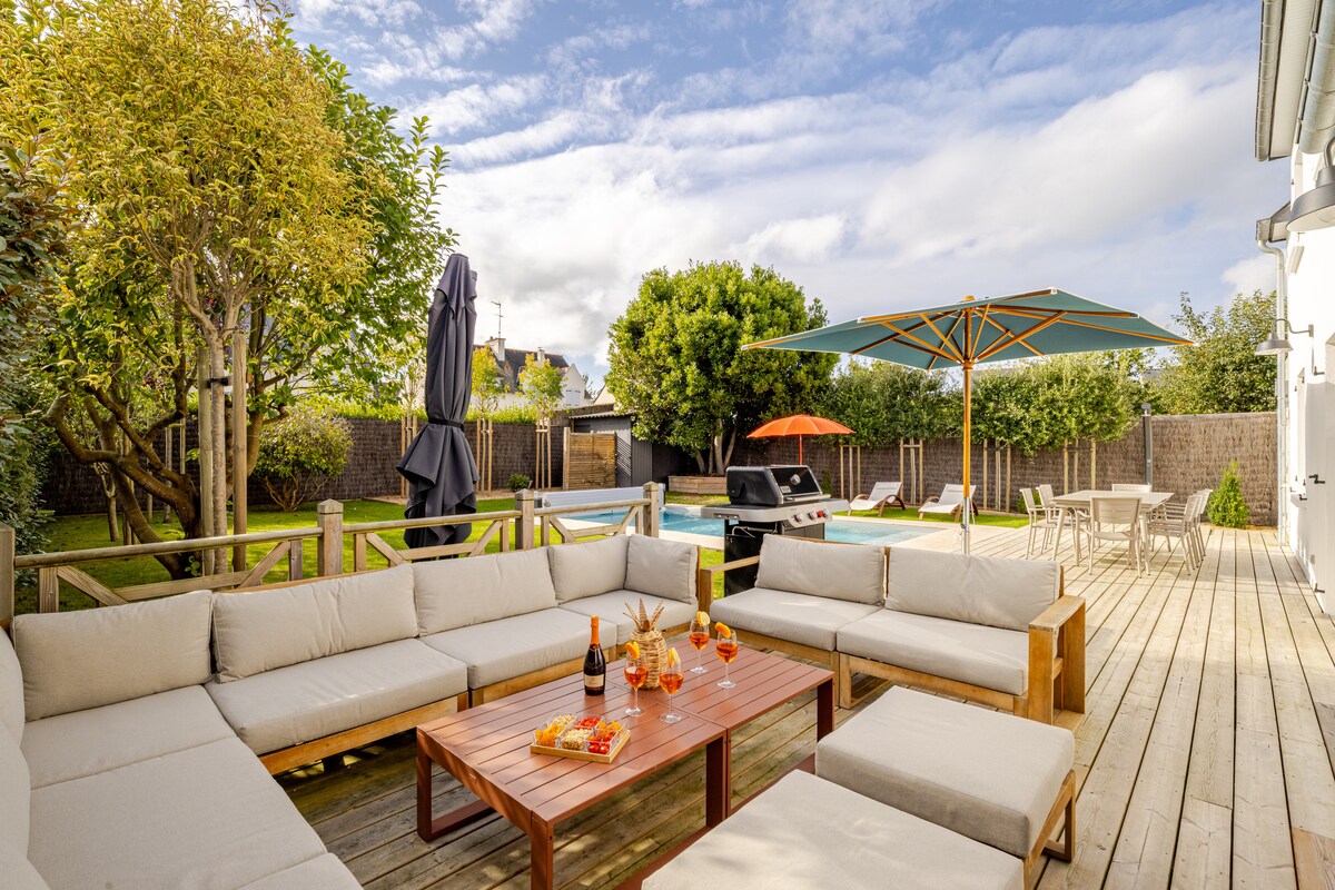 An outdoor seating area features a large sectional sofa arranged around a wooden coffee table, accompanied by refreshments. A shaded umbrella and colorful lounge chairs surround the private pool, while greenery adds to the serene ambiance.