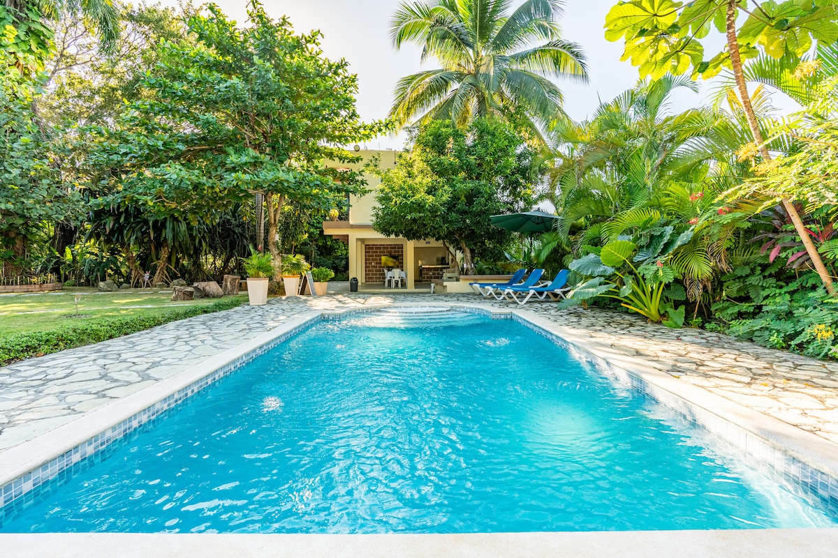 A serene swimming pool is surrounded by lush greenery and tall palm trees, creating a tranquil outdoor space. Lounge chairs are positioned near the water, while a shaded area includes seating for relaxation. The inviting blue water reflects the sunlight, enhancing the peaceful environment.