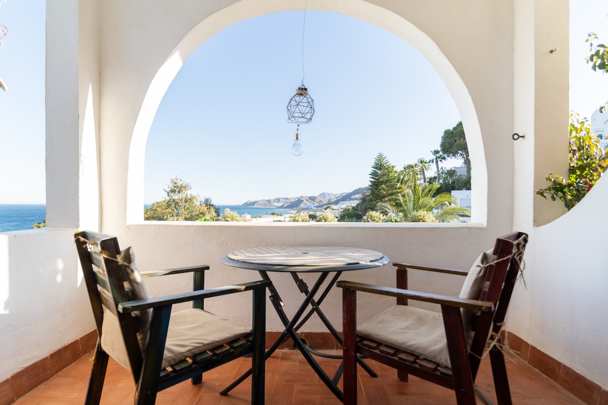 A balcony space features a round table surrounded by two wooden chairs. An arched window offers a view of the sea and lush greenery, while a hanging light fixture adds a simple decorative touch. The area is bright and open.
