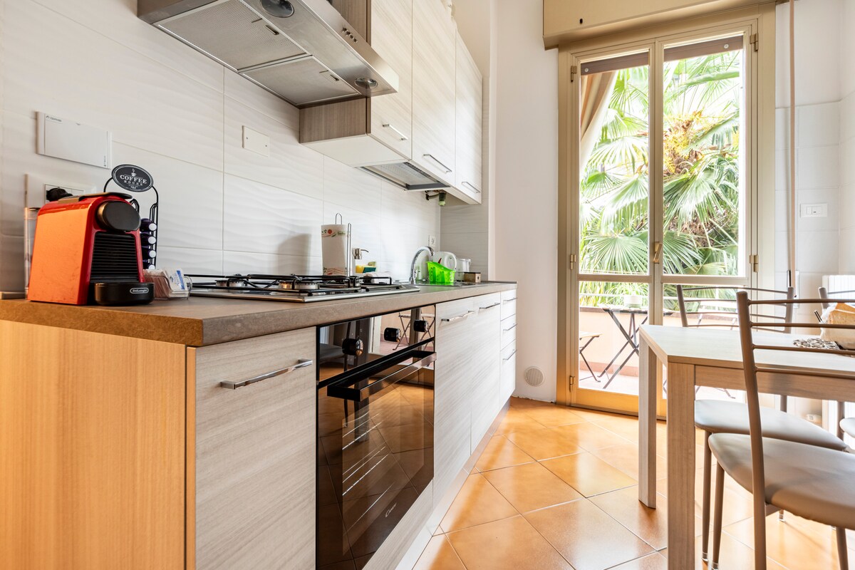 A modern kitchen is equipped with sleek cabinetry and stainless-steel appliances. A gas stove and an oven are integrated into the design. Natural light enters through large windows, illuminating the space. A small dining table is set for two, with views of the balcony's greenery.
