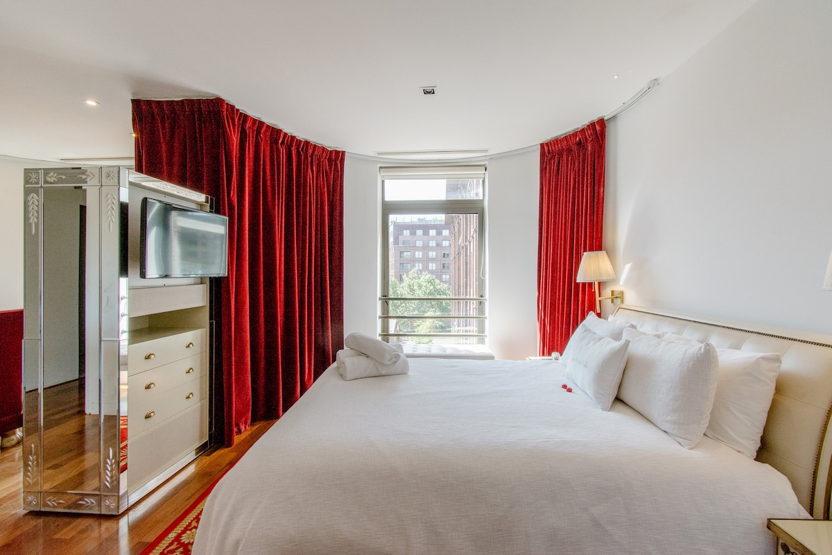 A spacious bedroom features a large bed with plush white bedding. Red drapes frame a window that allows natural light to brighten the room. A stylish dresser and a flat-screen TV are positioned nearby, creating a comfortable and inviting layout.