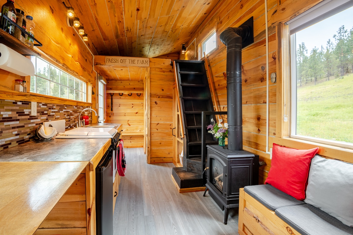 The interior features rustic blue pine walls and a compact kitchen with modern appliances. A small propane stove is positioned next to a comfortable seating area, where bright cushions add subtle color. A staircase leads to a loft area, with natural light filling the space through large windows.