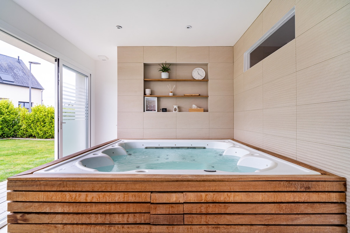 A modern jacuzzi is situated in a stylish room, framed by wooden paneling. Light enters through large glass doors, revealing a well-kept garden beyond. Neutral tones enhance the calming ambiance, while minimal decor, including a clock and plant, adorns the wall shelves.