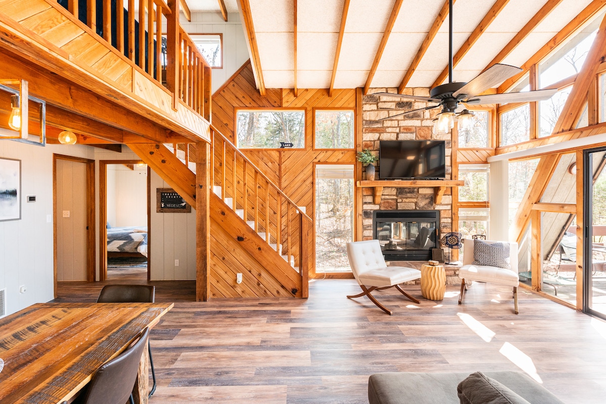 An open living space is highlighted by vaulted wooden ceilings and large windows allowing natural light to flood in. A stone fireplace acts as a focal point, complemented by modern furniture. A staircase leads to a loft area, while polished wooden flooring adds warmth.