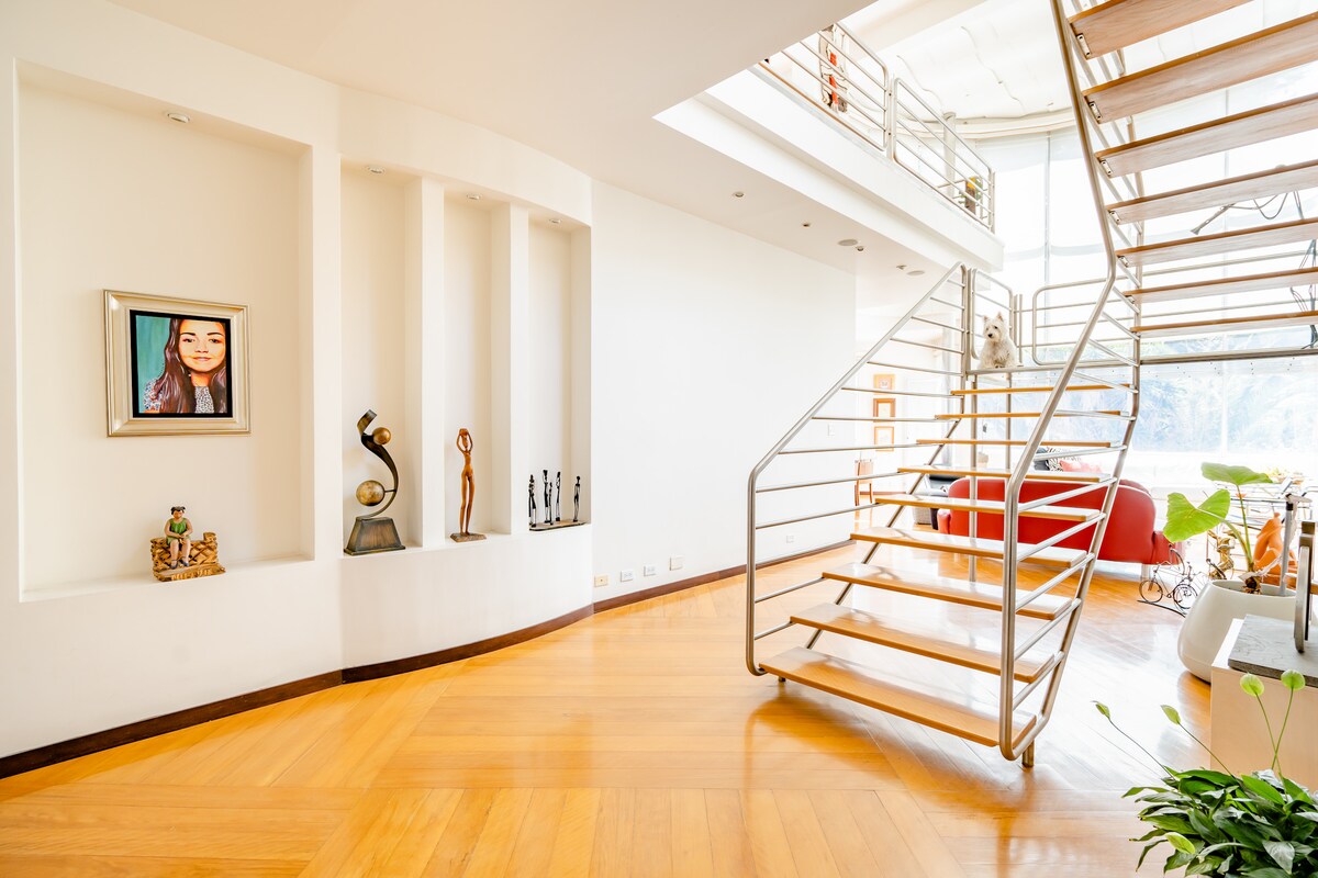 A well-lit entrance features a wooden staircase leading to the second floor. Decorative shelves display various art pieces, while a vibrant red chair provides an inviting seating option. Natural light fills the space through large windows, enhancing the open layout.