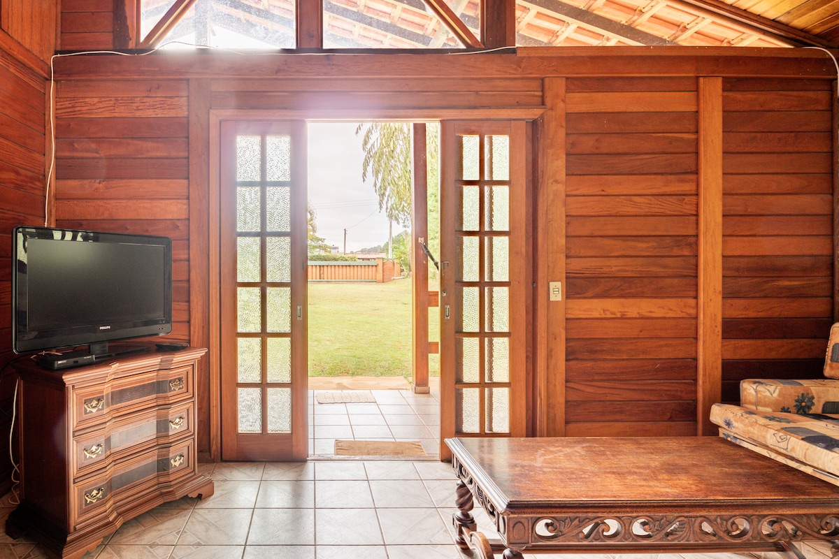 A wooden interior is highlighted by large glass doors leading to a grassy outdoor area. A dark wooden coffee table rests atop tiled flooring, complemented by a comfortable seating arrangement and a television on an ornate cabinet.