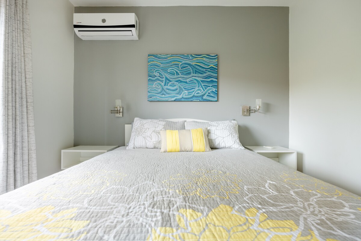 A spacious bedroom features a large bed adorned with a light grey quilt and yellow accents. Wall-mounted lamps provide gentle illumination beside each side of the bed. Above, a blue wave painting adds a serene touch, with a modern air conditioning unit mounted on the wall.