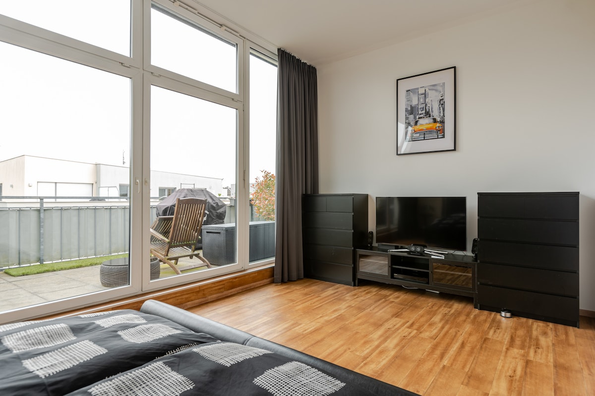 A modern room features large glass windows that extend to a private terrace. A black entertainment unit stands against the wall, with a television positioned on top. Light wood flooring complements the minimalist design throughout the space.