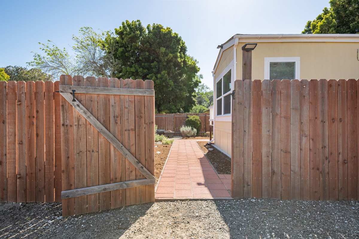 A wooden gate opens to a paved walkway leading towards a light-colored cottage. Lush greenery and landscaping are visible within the fully fenced yard, providing a private and inviting entrance to the property.