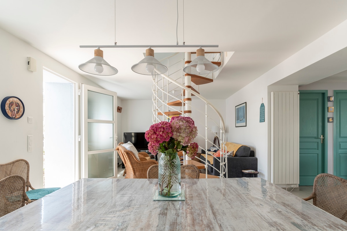 The dining area features a large table with a textured surface, surrounded by woven chairs. A vase with colorful flowers sits at the center. A spiral staircase leads to an upper level, and natural light fills the space through large doors.