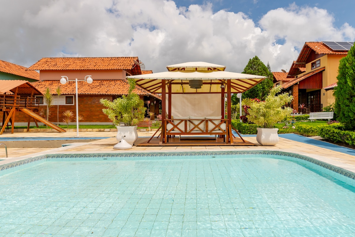 A well-maintained outdoor space features a clear pool surrounded by manicured gardens. A wooden gazebo is positioned near the pool, offering shaded seating. Nearby, several charming chalets with terracotta roofs can be seen against a backdrop of blue sky and fluffy white clouds.