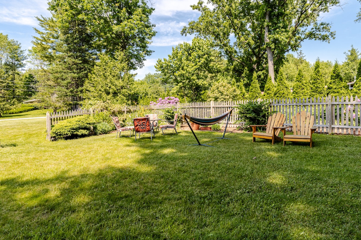A spacious yard is outlined by a wooden fence, featuring a cozy seating area with Adirondack chairs and a hammock. Lush green grass is surrounded by trees and colorful flowers, creating a serene outdoor space ideal for relaxation.