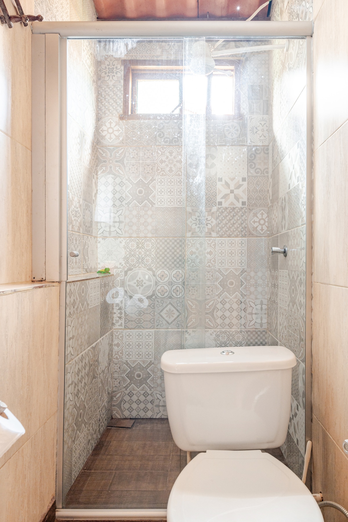 A compact bathroom is presented, featuring a glass-enclosed shower with decorative tile patterns. A modern toilet is positioned beside the shower, with natural light filtering in from a small upper window.