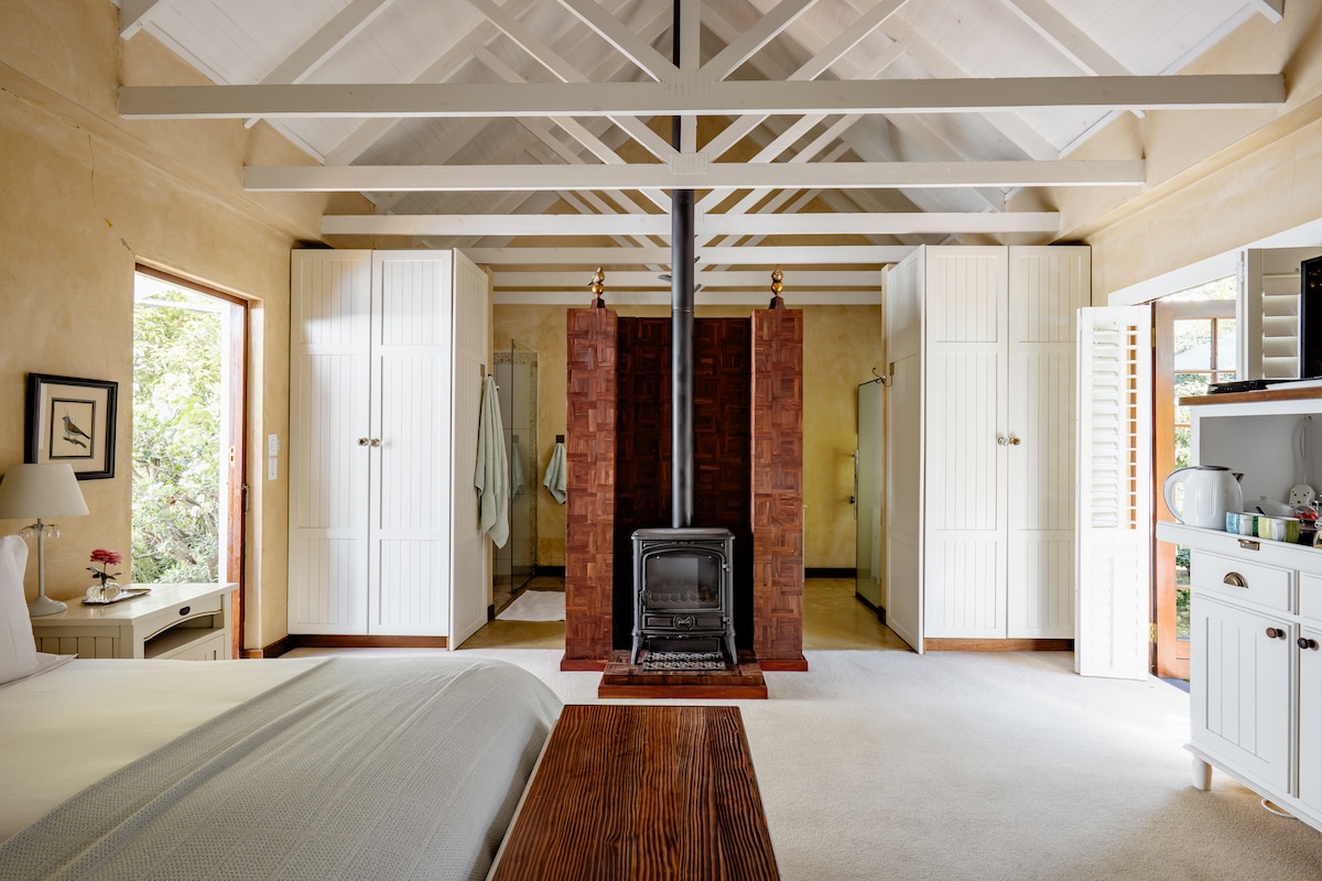 A spacious guest suite featuring a high ceiling with wooden beams and light-colored walls. Two windows allow natural light to fill the room, while a cozy gas fireplace serves as a focal point. Open cupboards and a kitchenette are visible, enhancing convenience.
