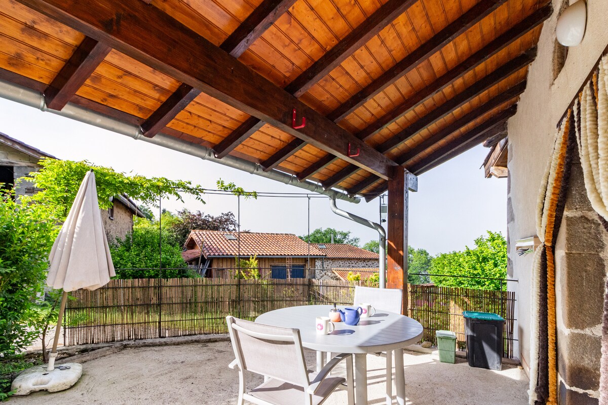 A covered outdoor terrace features a round table with multiple chairs and a sun umbrella. A rustic wooden beam ceiling adds character. Lush greenery is visible in the background, complementing the serene countryside setting.