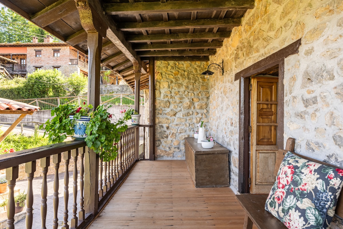 The covered porch displays a traditional wooden railing, featuring potted flowers hanging from its sides. A simple wooden bench is positioned alongside, while a decorative table rests against the wall. Natural light highlights the stone facade of the house and the surrounding greenery.