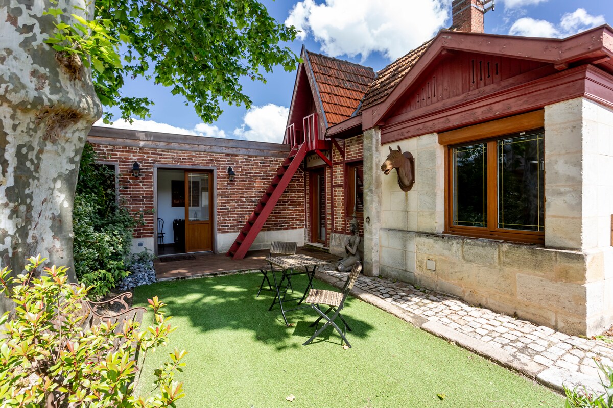 A charming exterior of the guest house displays a mix of brick and stone walls, framed by lush greenery. A seating area with two metal chairs and a small table is situated on manicured grass. Large windows allow natural light to brighten the space.