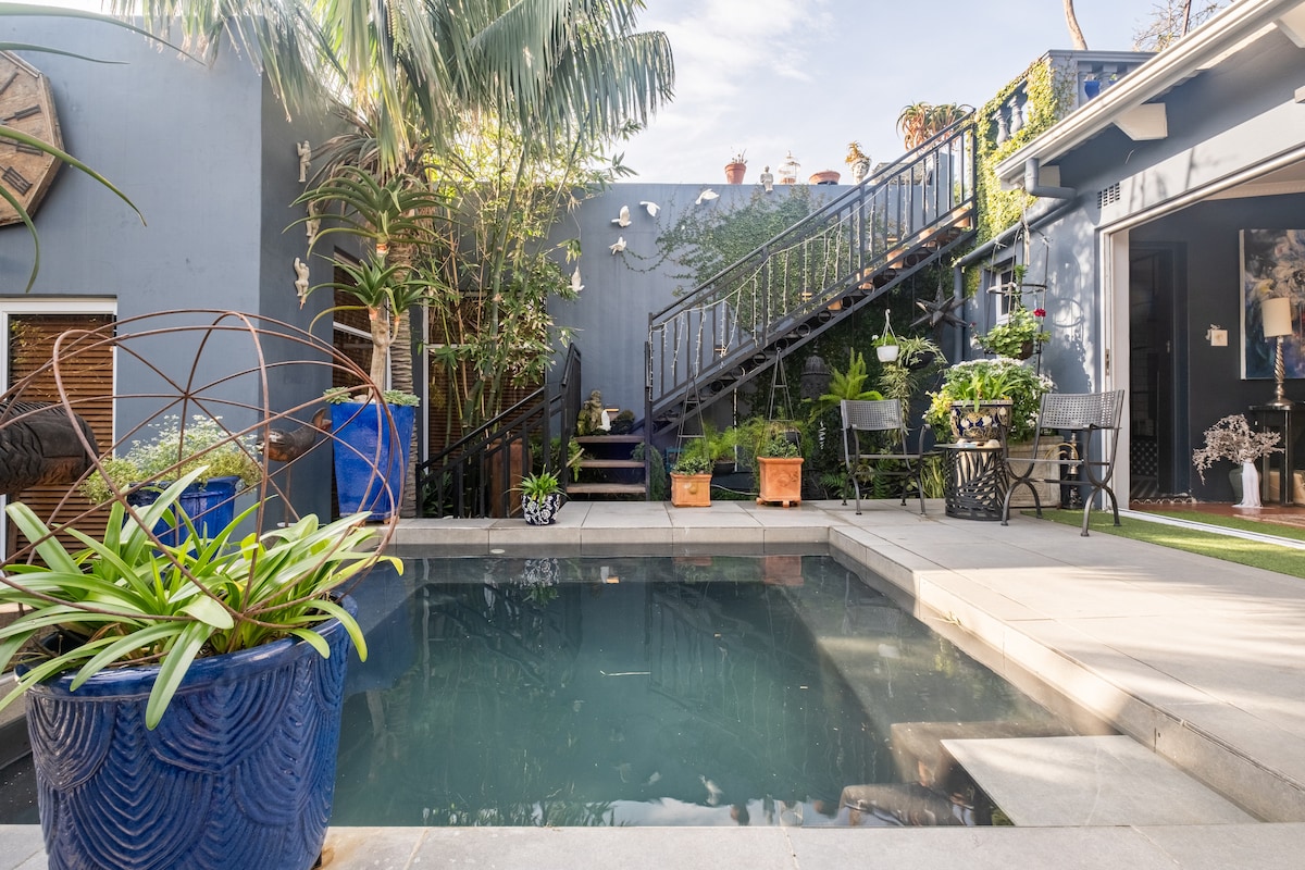A tranquil garden area features a salt-water cocktail pool surrounded by lush greenery and decorative pots. A staircase lined with plants leads to an upper level, while seating is available nearby for relaxation. Natural light enhances the serene atmosphere.