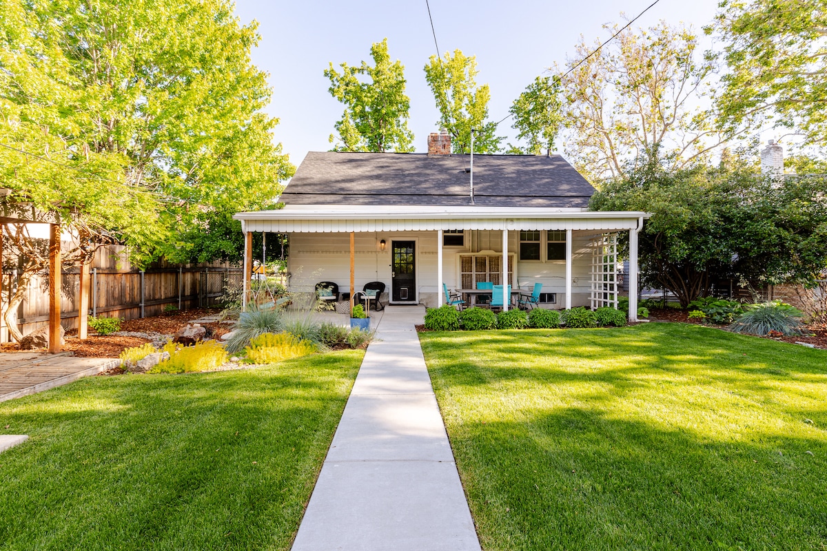 A well-maintained exterior of a home is featured, highlighted by a wide, inviting pathway leading to a covered porch. Lush green grass surrounds vibrant flower beds, while trees provide shade, creating a serene environment. Outdoor seating is arranged on the porch, inviting relaxation.