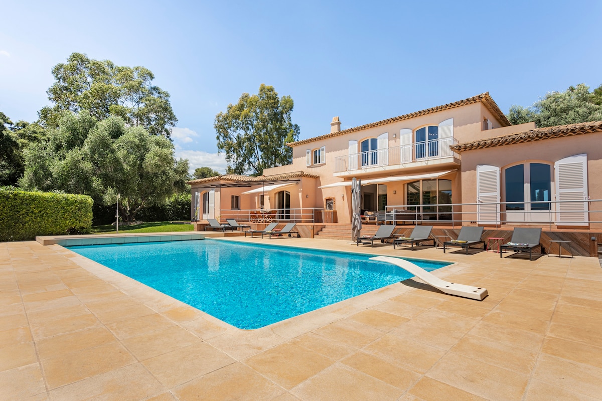A spacious villa is showcased with a large swimming pool surrounded by sun loungers. The exterior features terracotta tiles and light-colored walls, with numerous windows allowing ample natural light. Lush greenery frames the property, enhancing the inviting outdoor space.