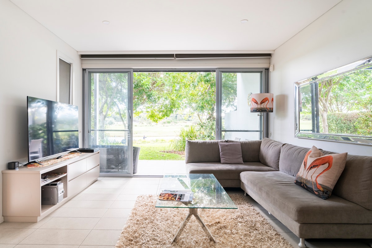 A modern living area features a spacious sectional sofa and a glass coffee table placed on a soft area rug. Large sliding glass doors provide views of the lush greenery outside, allowing natural light to fill the room. A sleek television is set against the wall.